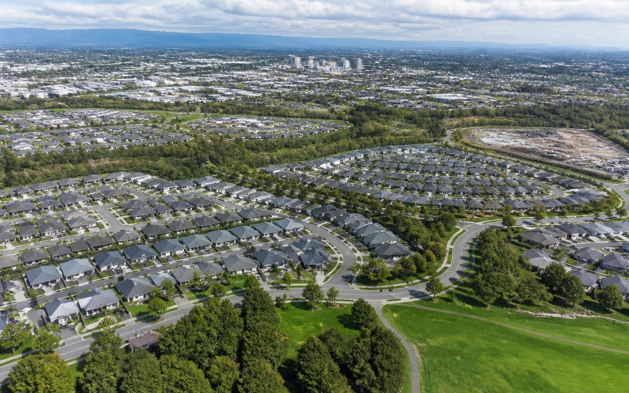 Elevated daylight view of a planned neighbourhood with curving residential streets, green belts and a town centre in the distance.